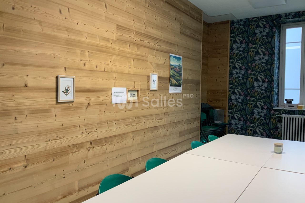 Mur en bois clair décoré de cadres, table blanche entourée de chaises vertes, et mur floral en arrière-plan.
