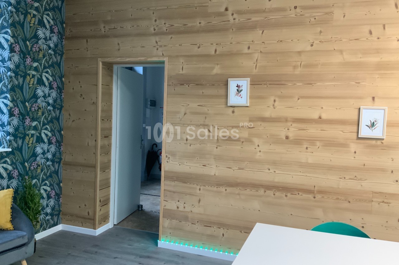 Mur en bois clair avec deux cadres décoratifs, porte ouverte sur un couloir, sol en parquet et coin de mobilier visible.