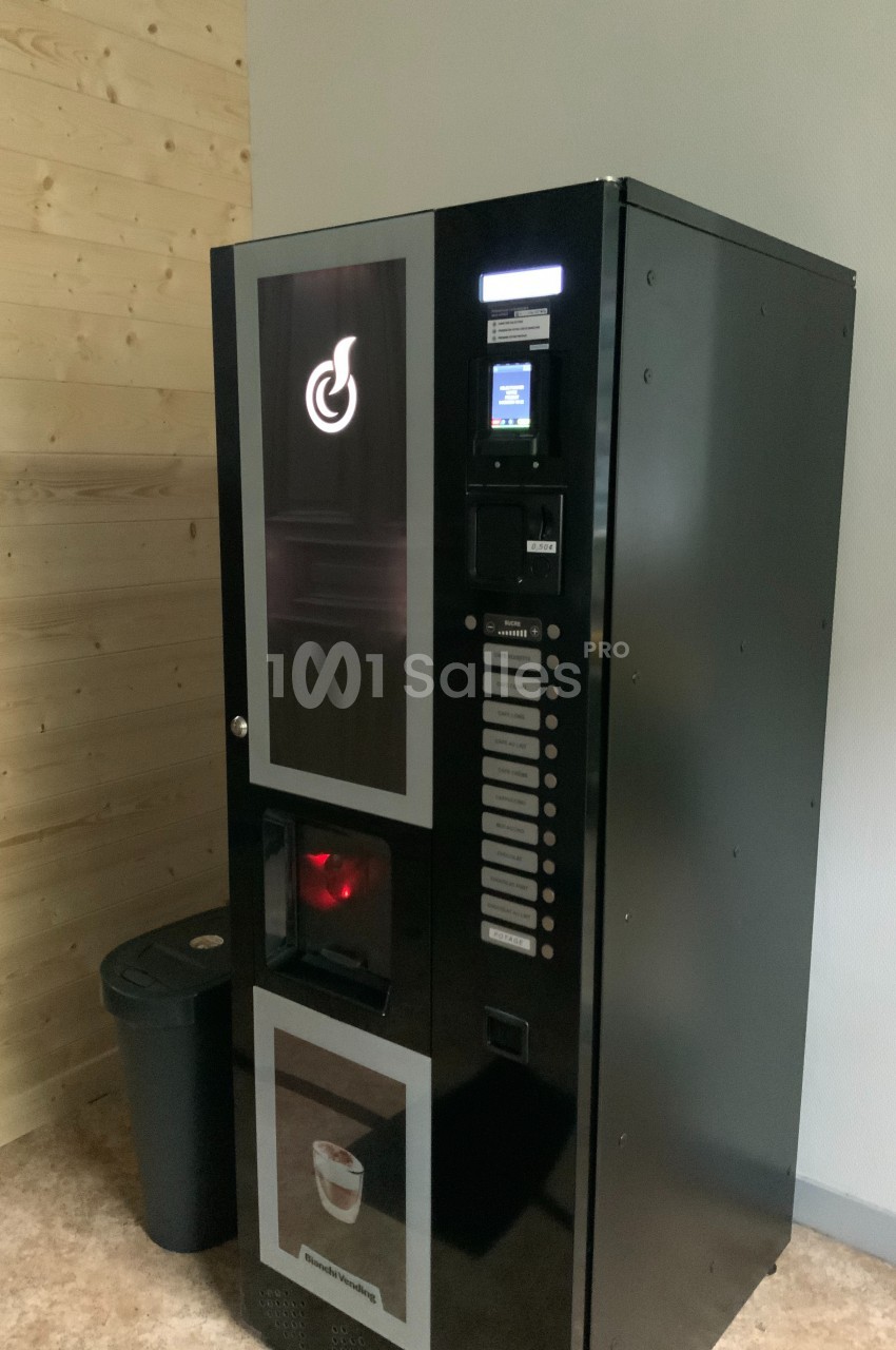 Distributeur automatique de boissons chaudes noir, installé près d'un mur en bois clair et d'une poubelle noire.