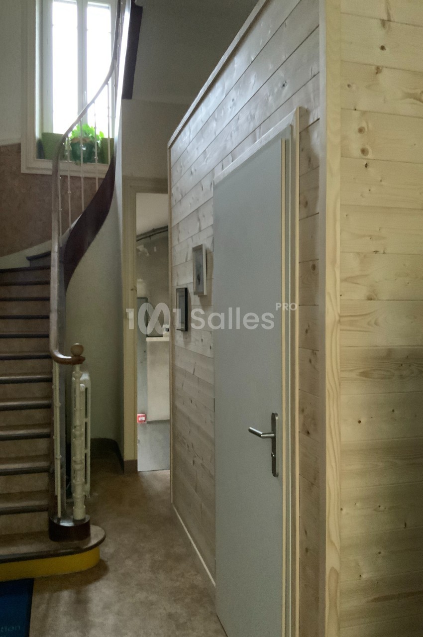 Escalier en bois courbé à côté d'une cabine en bois clair avec une porte blanche, dans un intérieur lumineux.