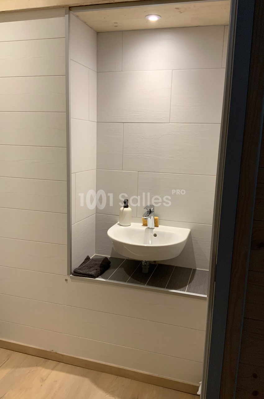 Lavabo blanc suspendu dans une alcôve carrelée, éclairée par un spot, avec accessoires et serviette posée à côté.
