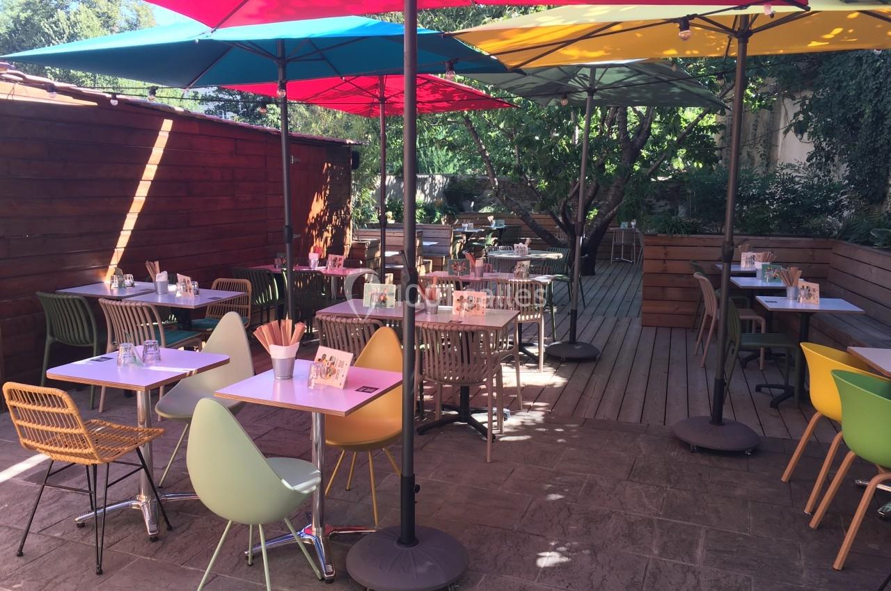 Terrasse extérieure avec tables et chaises colorées sous des parasols, entourée de verdure et de murs en bois.