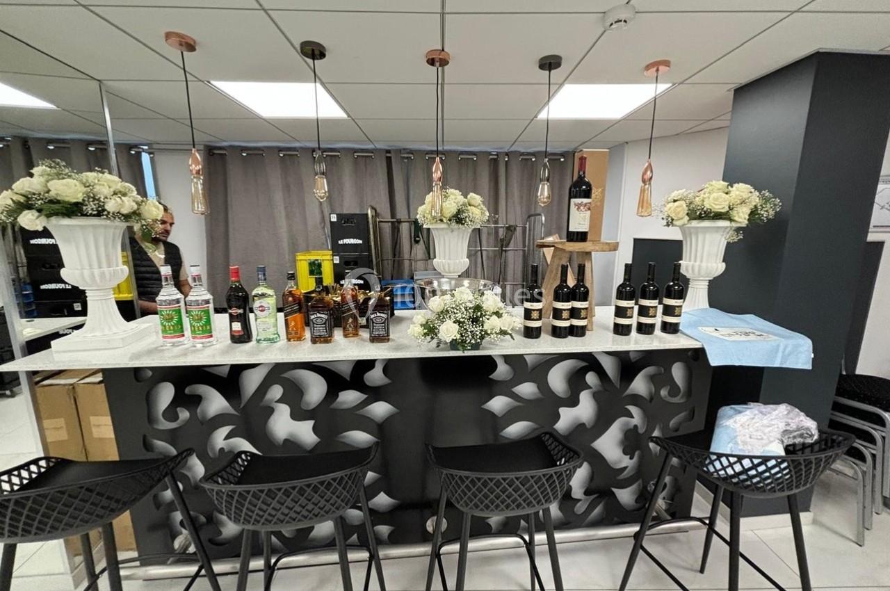 Bar décoré avec des fleurs blanches, bouteilles d'alcool alignées sur le comptoir et tabourets noirs devant.