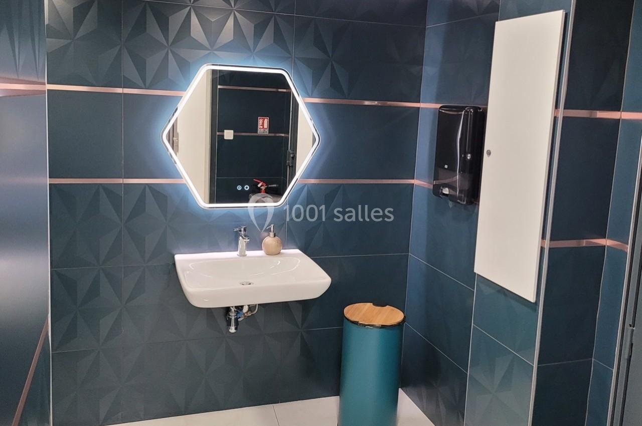 Lavabo blanc avec miroir rétroéclairé hexagonal, mur en carrelage bleu texturé et poubelle métallique turquoise.