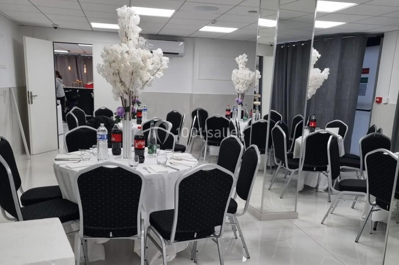 Salle de réception avec tables rondes dressées, chaises noires et décorations florales blanches.