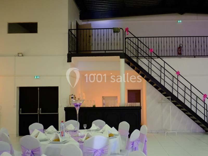 Salle de réception décorée avec des chaises blanches ornées de nœuds roses, un escalier et un bar en arrière-plan.