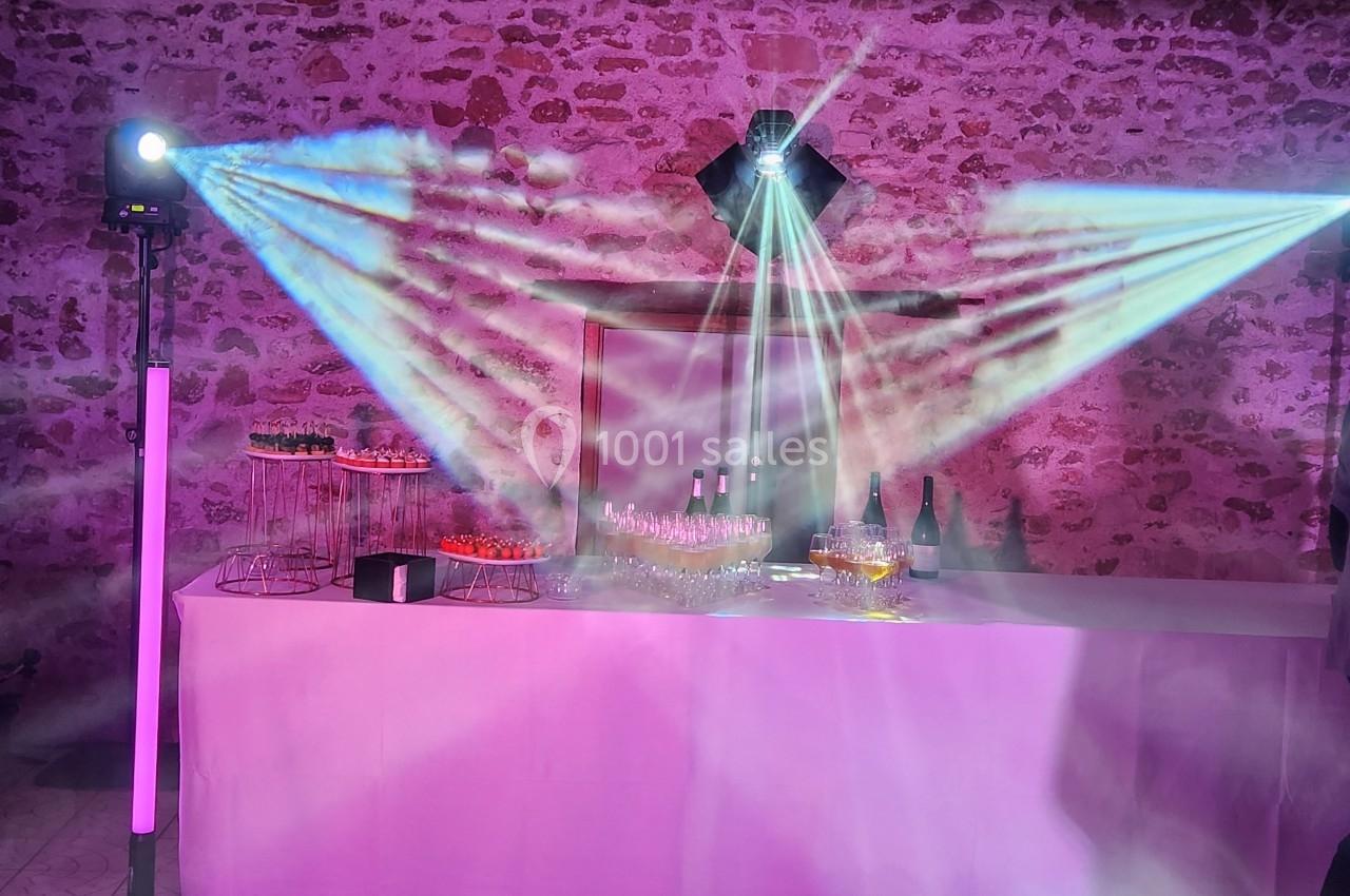 Table avec des verres, bouteilles et amuse-bouches, éclairée par des projecteurs colorés dans une salle en pierre.