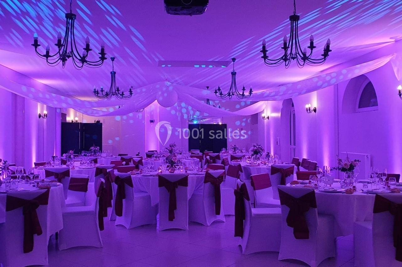 Salle de réception décorée avec des tables rondes, nappes blanches, chaises ornées de nœuds rouges et éclairage violet.