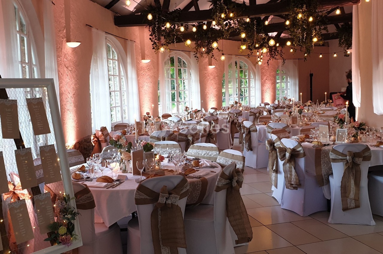 Salle de réception décorée avec des tables dressées, des chaises ornées de rubans et un plan de table à l'entrée.