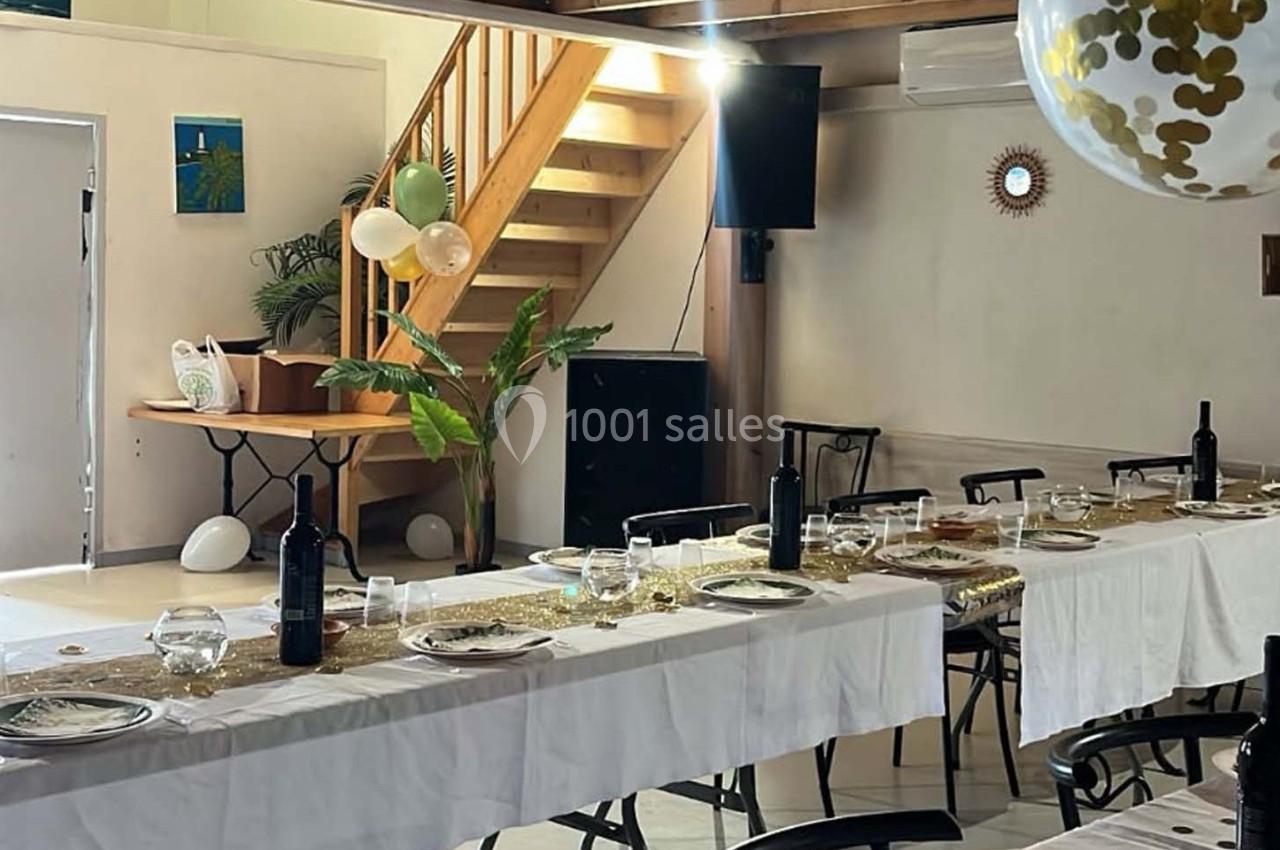 Salle décorée pour un événement, avec tables dressées, nappes blanches, bouteilles de vin et ballons suspendus.