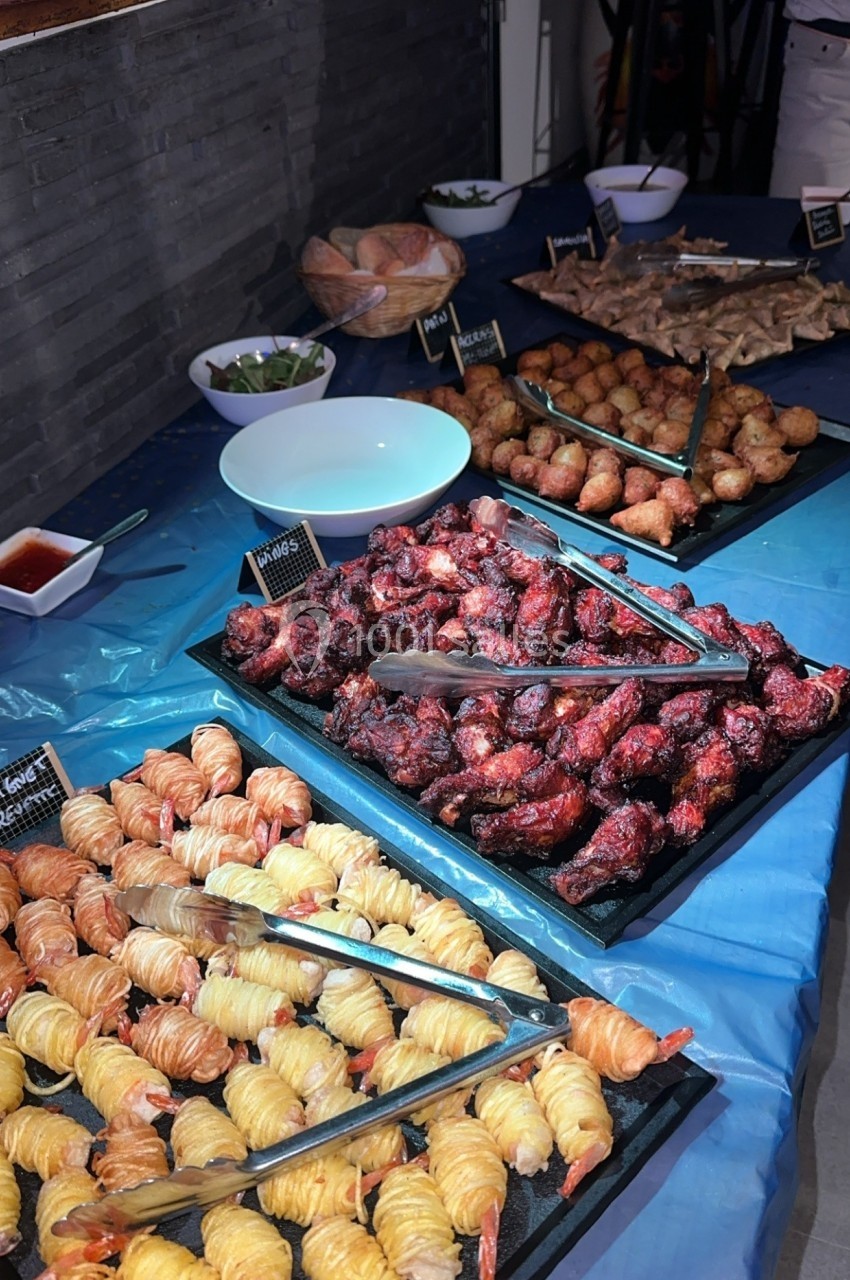 Buffet avec divers plats, dont crevettes enroulées, ailes de poulet grillées et boulettes, disposés sur une nappe bleue.