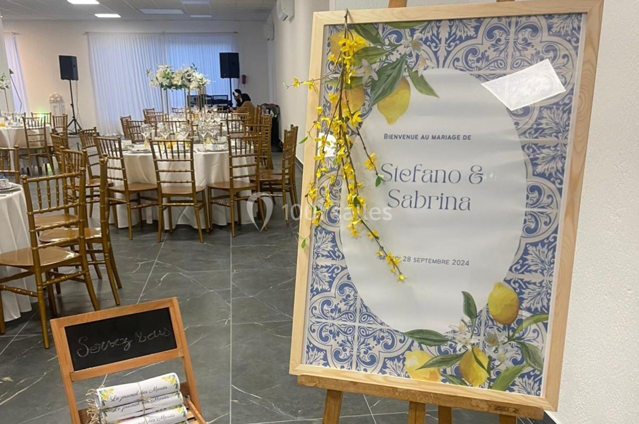 Panneau de bienvenue décoré pour un mariage, placé dans une salle avec tables dressées et chaises en bois.