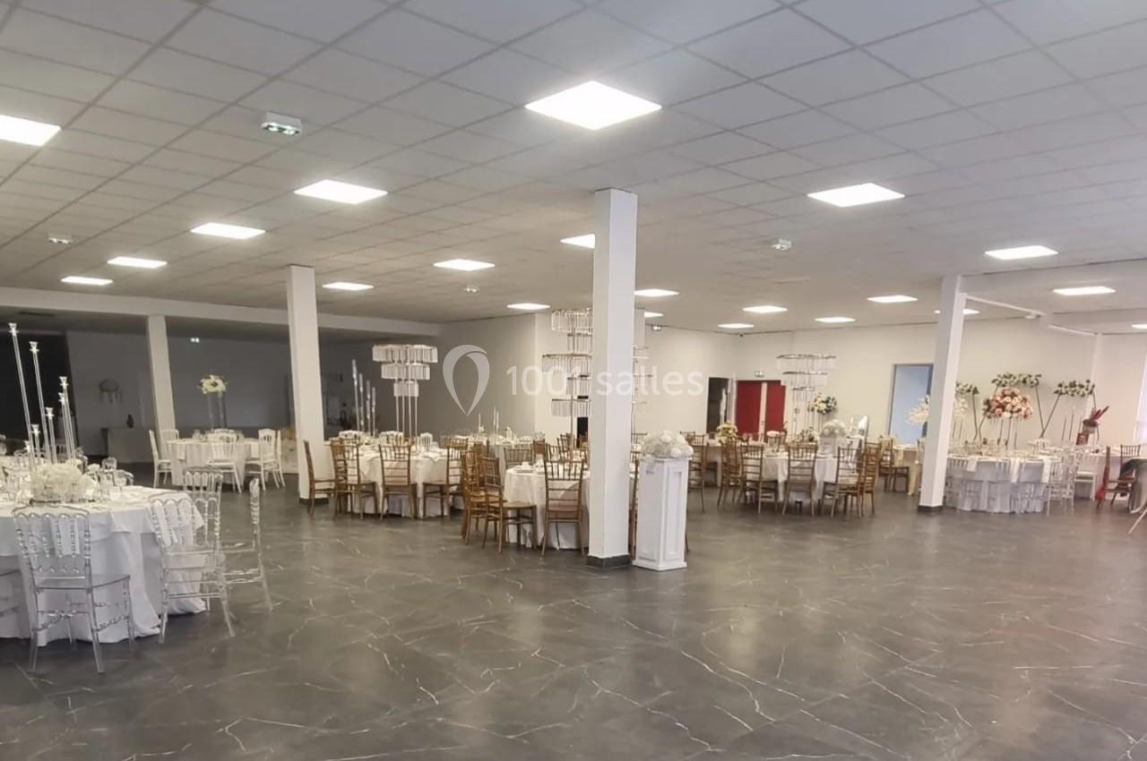 Salle de réception spacieuse avec tables décorées, chaises dorées et blanches, éclairage au plafond.