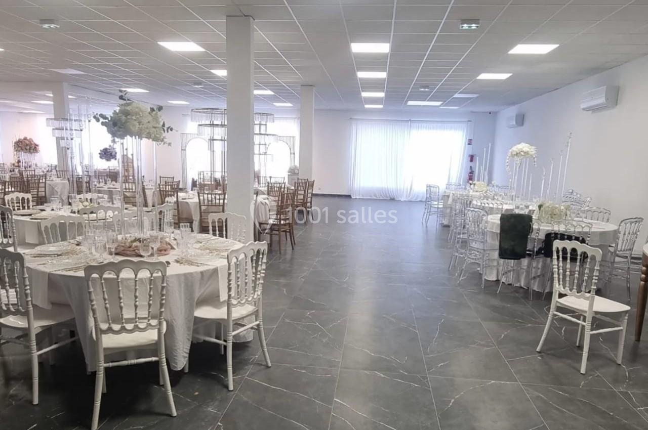 Salle de réception lumineuse avec tables rondes dressées, chaises blanches et décorations florales élégantes.