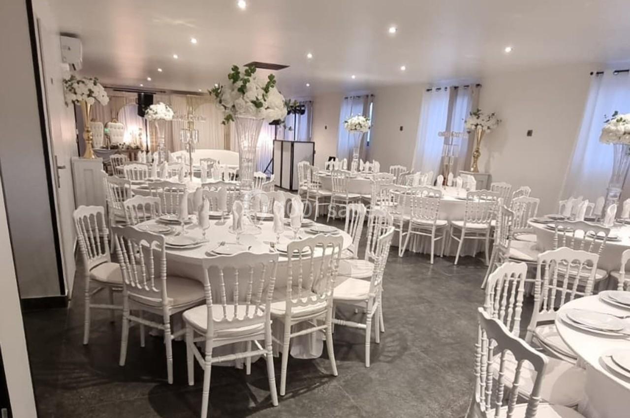 Salle de réception décorée avec des tables rondes, nappes blanches, chaises élégantes et arrangements floraux.