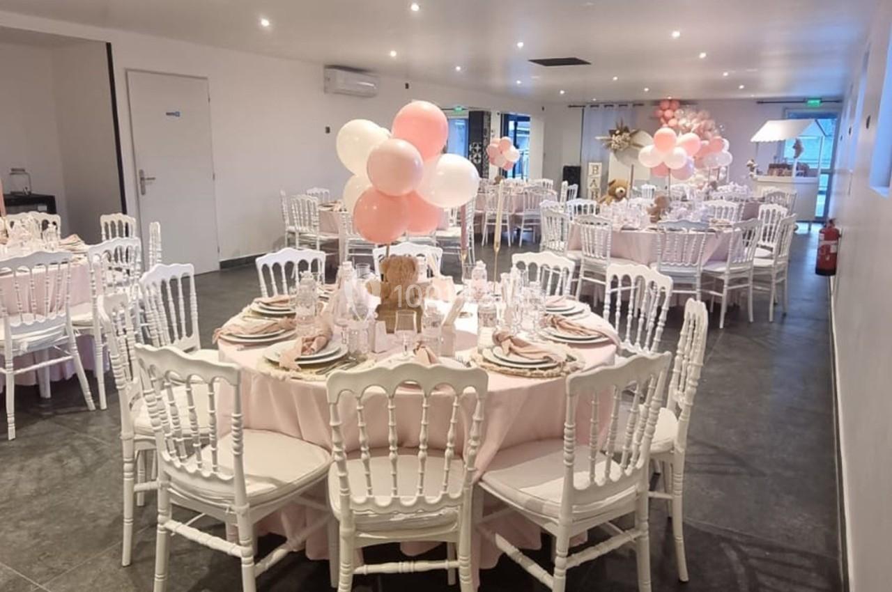 Salle décorée pour un événement avec des tables rondes, nappes roses, chaises blanches et ballons roses et blancs.