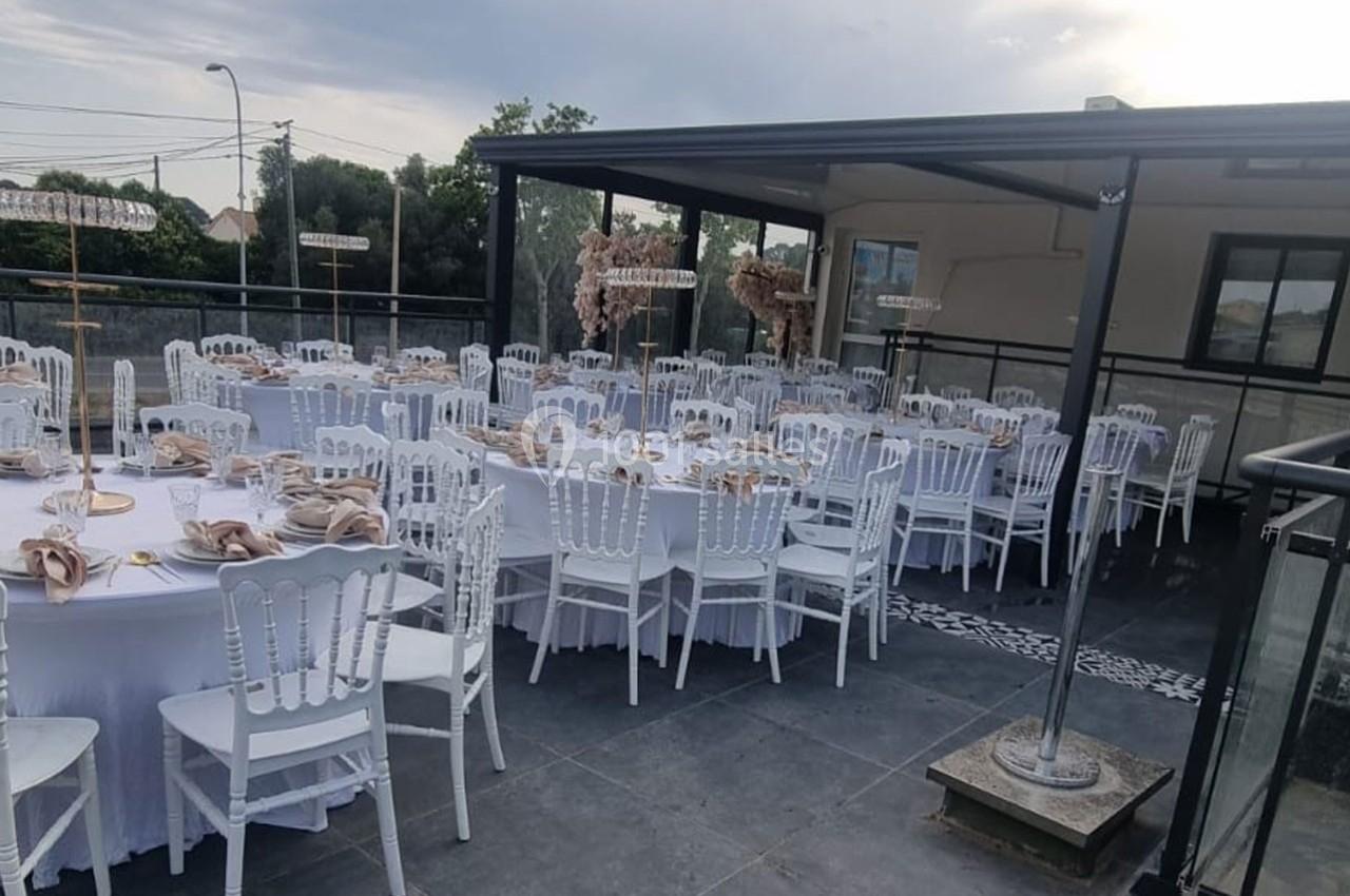 Terrasse aménagée pour un événement avec des tables rondes dressées, nappes blanches et chaises blanches élégantes.