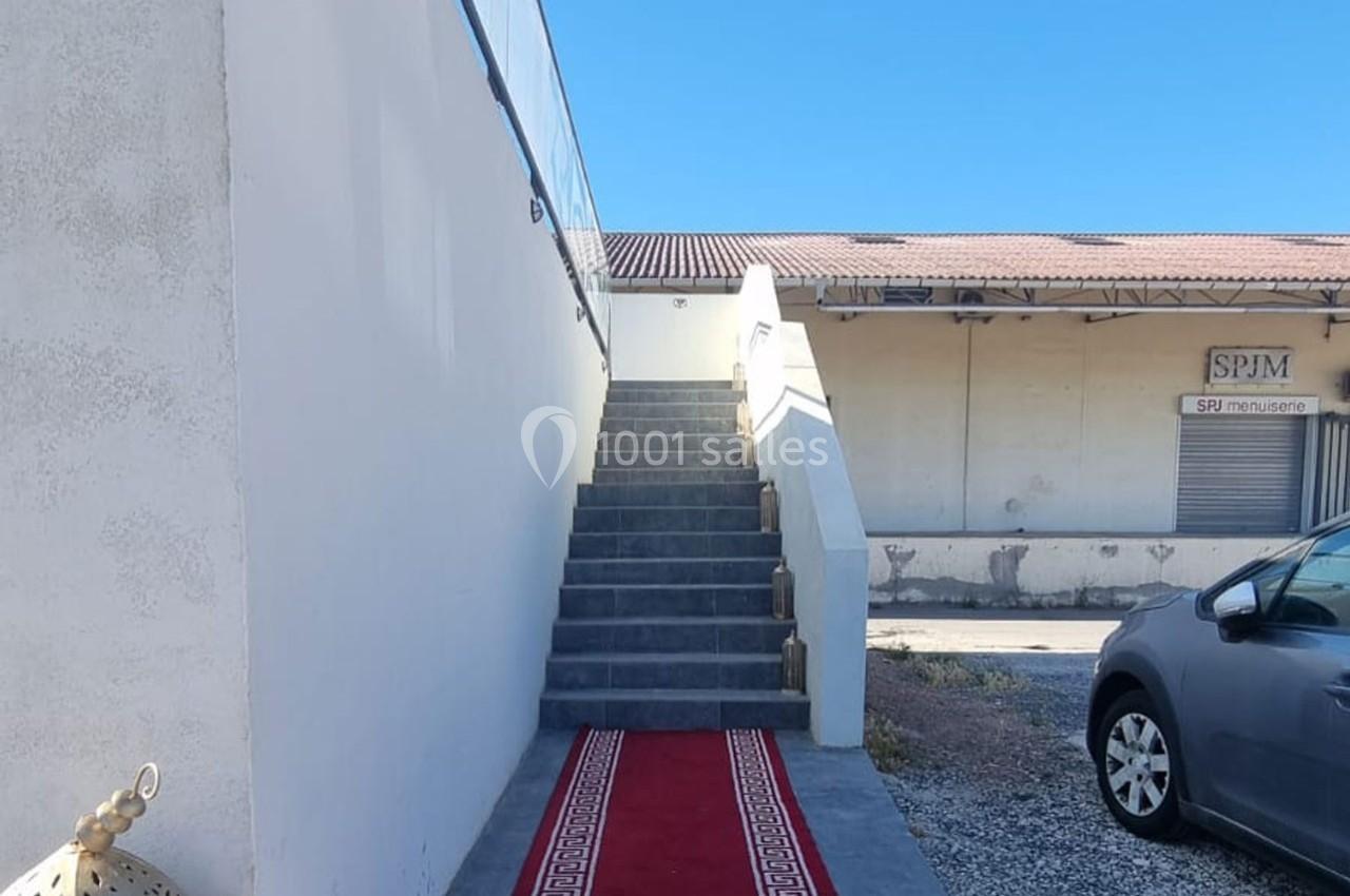 Escalier extérieur en béton avec tapis rouge, menant à une terrasse, près d'un bâtiment industriel et d'une voiture grise.