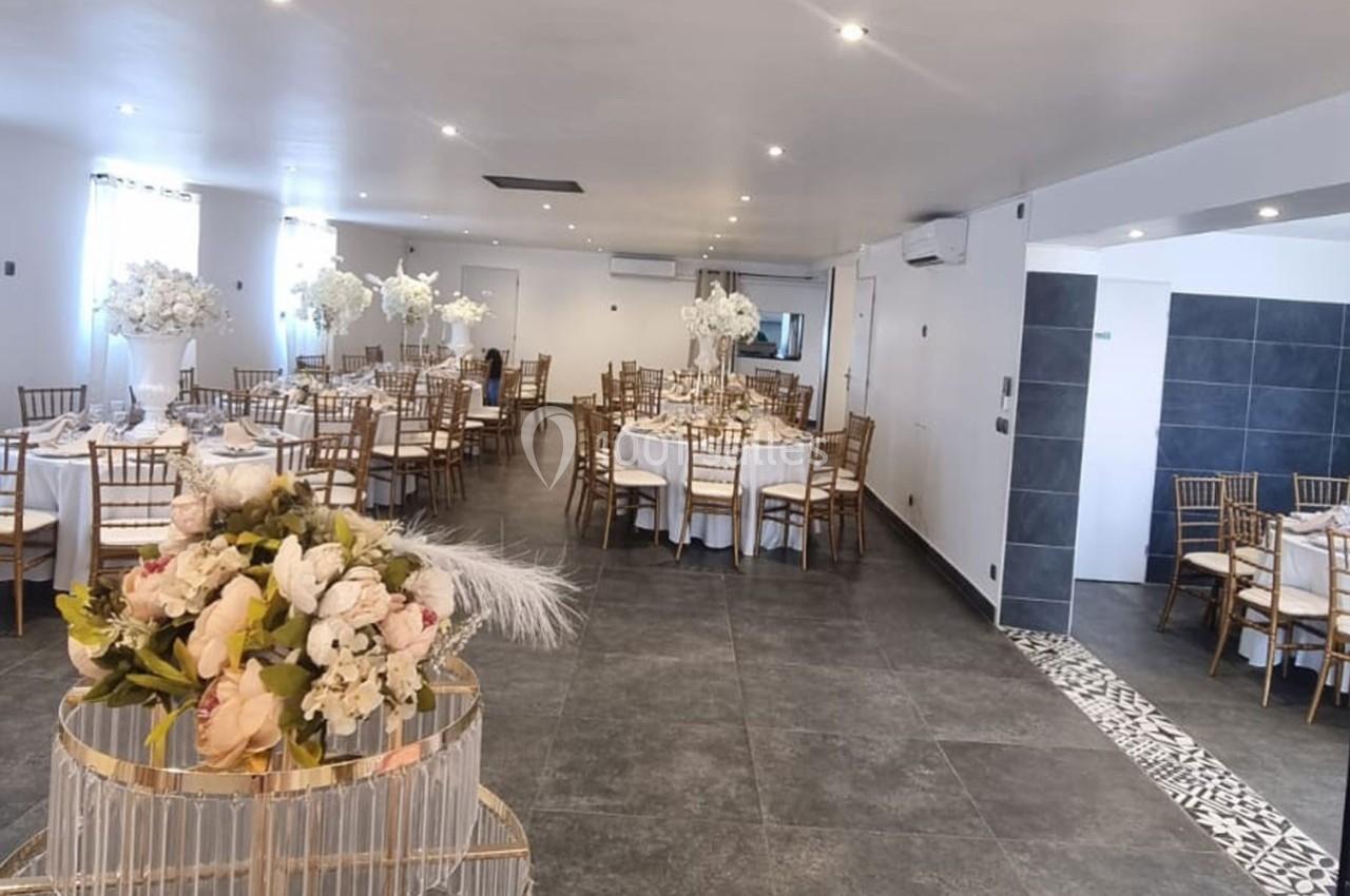 Salle de réception lumineuse avec tables rondes dressées, chaises dorées et décorations florales élégantes.