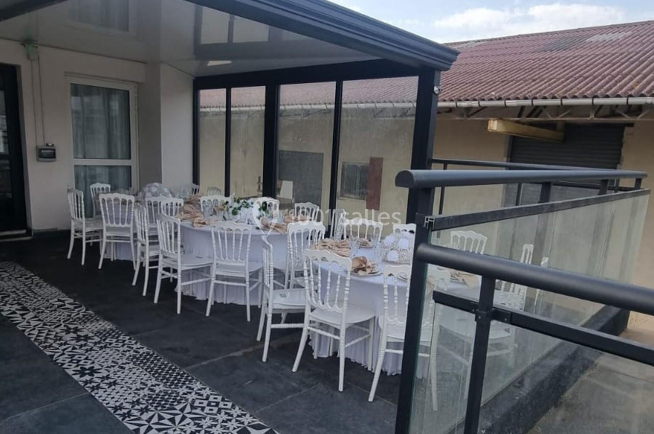 Tables dressées avec nappes blanches et chaises élégantes sur une terrasse couverte, prêtes pour un repas ou un événement.