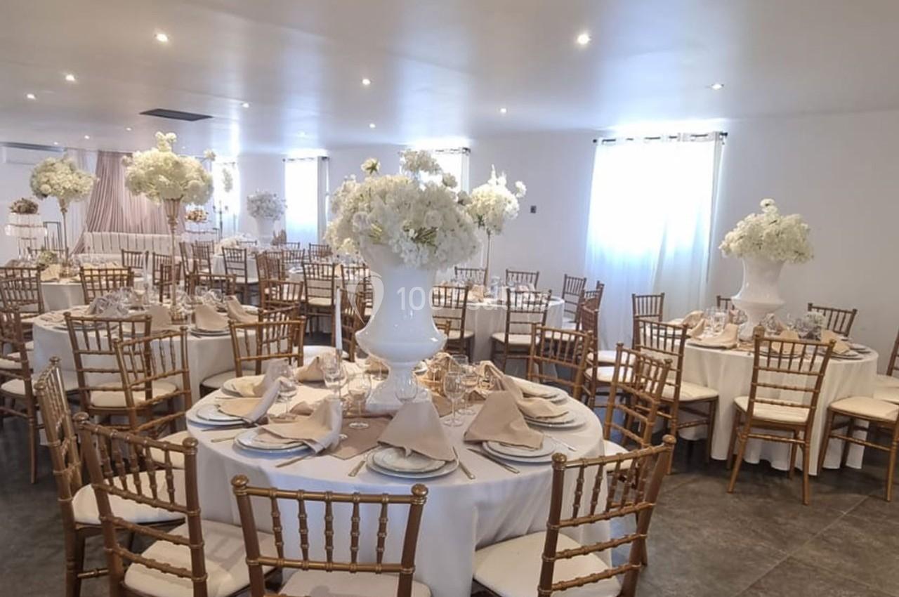 Salle de réception décorée avec des tables rondes dressées, nappes blanches, chaises dorées et centres de table fleuris.