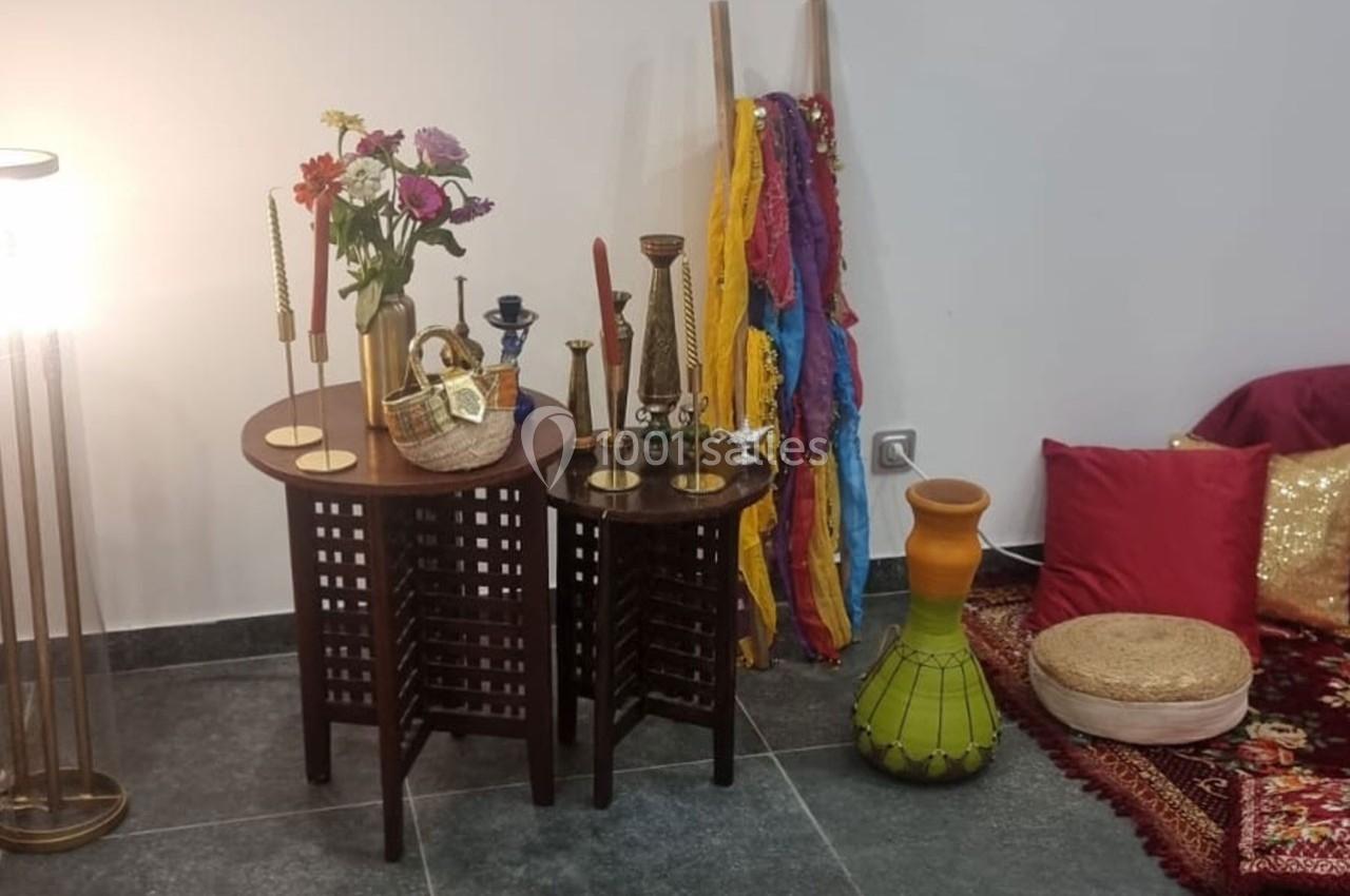 Table d'appoint en bois avec bougeoirs, fleurs et paniers, entourée de coussins, tissus colorés et un vase décoratif.