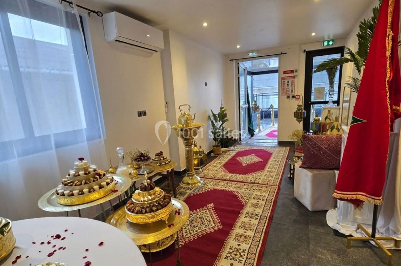 Salle décorée pour une réception avec tapis rouges, plateaux de pâtisseries dorés et drapeau marocain.