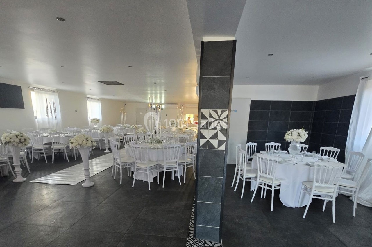 Salle de réception élégamment décorée avec des tables rondes, nappes blanches, chaises blanches et arrangements floraux.