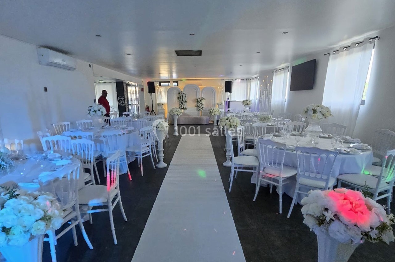 Salle de réception décorée avec des tables rondes, des chaises blanches et une allée centrale bordée de fleurs blanches.
