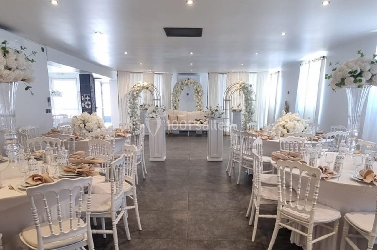 Salle de réception décorée en blanc avec des tables rondes, des chaises élégantes et des arches fleuries.