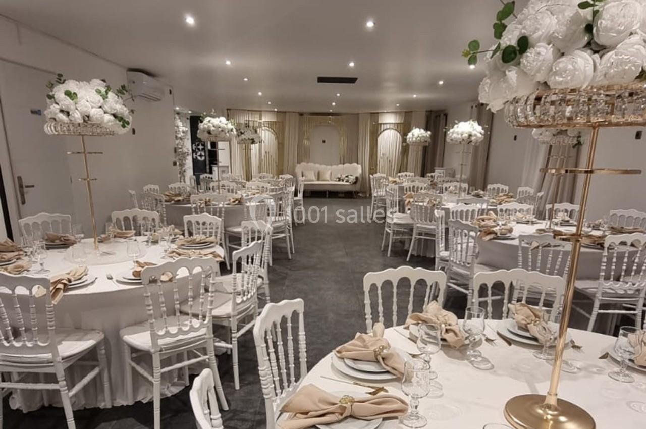 Salle de réception élégamment décorée avec des tables rondes, nappes blanches, chaises blanches et centres de table fleuris.
