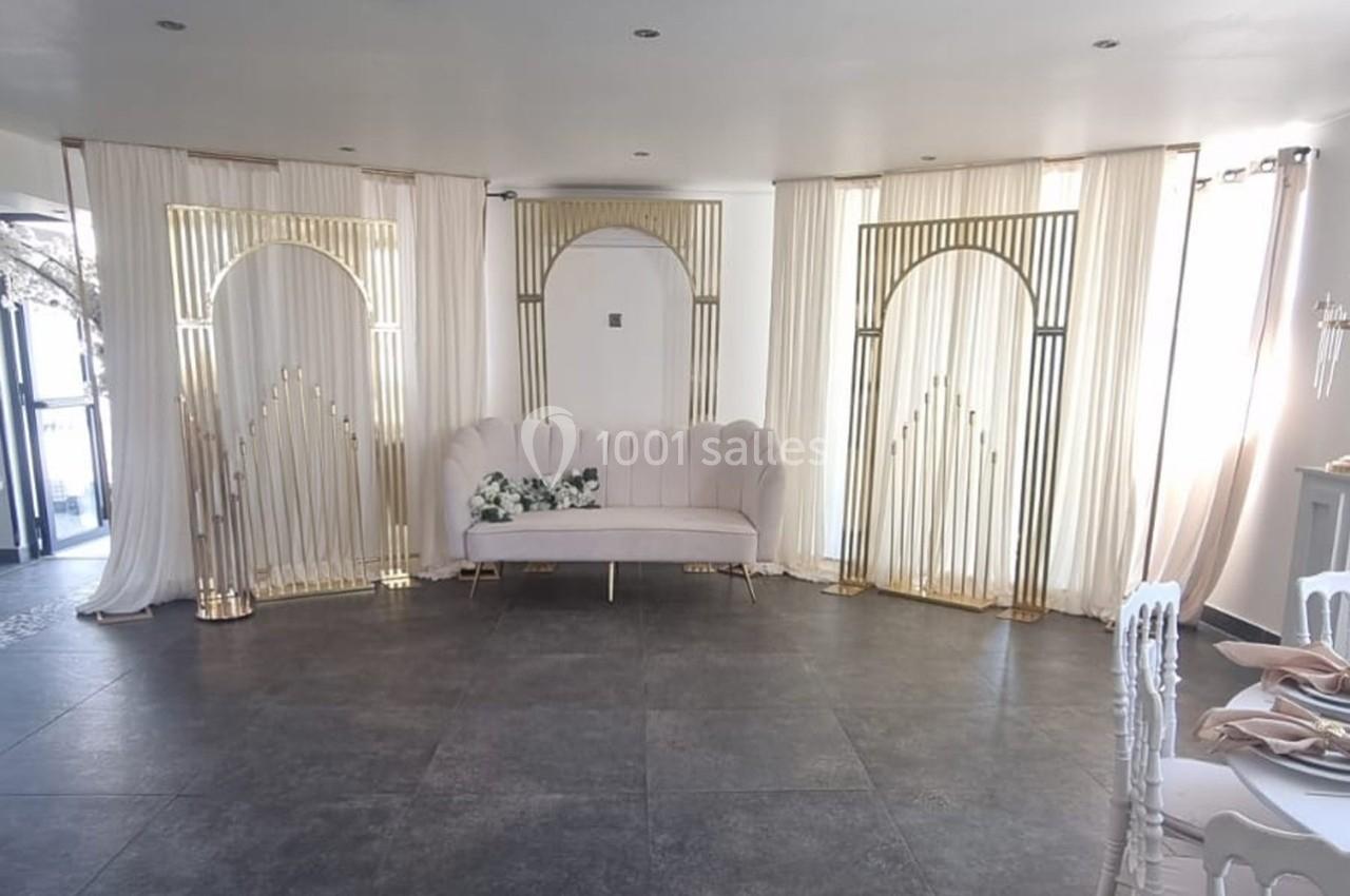 Canapé beige entouré de structures dorées et rideaux blancs dans une salle lumineuse au sol en carrelage gris.
