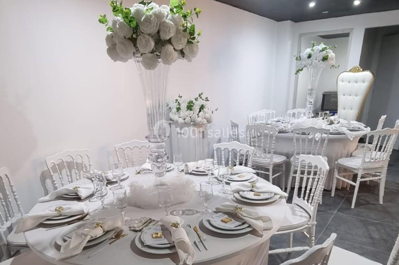 Salle décorée pour un événement avec tables rondes, nappes blanches, chaises assorties et centres de table fleuris.