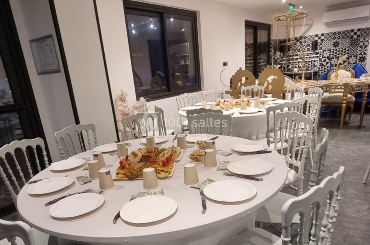 Salle décorée avec des tables rondes dressées, comprenant assiettes, couverts, gobelets et plats au centre.