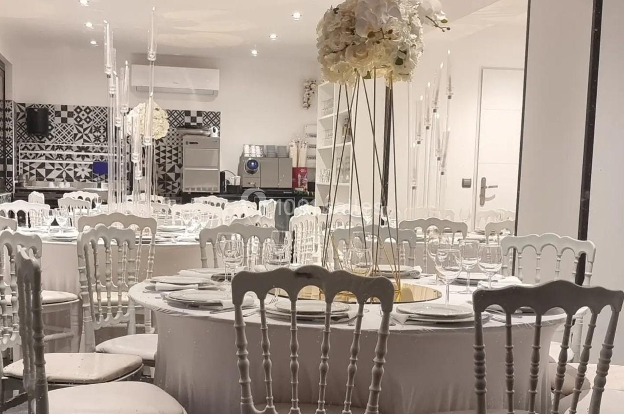 Salle de réception élégante avec tables rondes dressées, chaises blanches et centre de table floral.