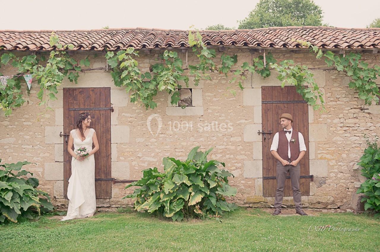 Une mariée en robe blanche et un marié en tenue décontractée posent devant un mur en pierre avec des portes en bois.