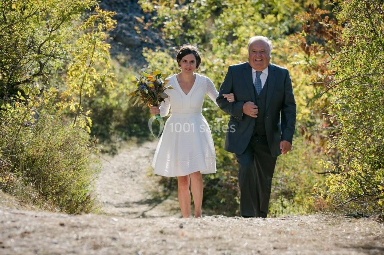 Une femme en robe blanche tenant un bouquet marche aux côtés d'un homme en costume dans un chemin entouré de végétation.