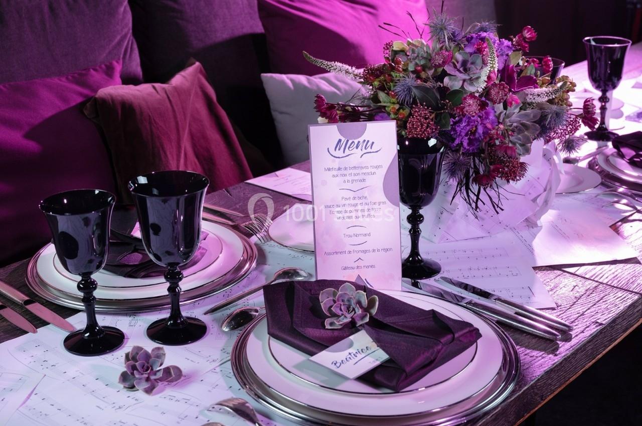 Table élégamment dressée avec vaisselle noire, serviettes violettes, menu imprimé et bouquet floral coloré.