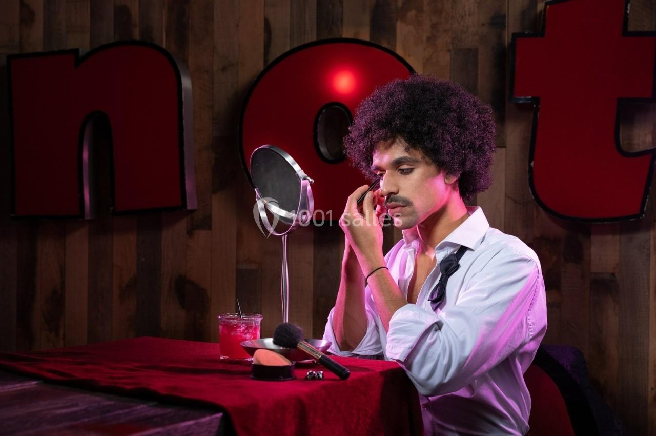 Un homme maquillé, assis devant un miroir, applique du maquillage sur son visage dans un décor éclairé.