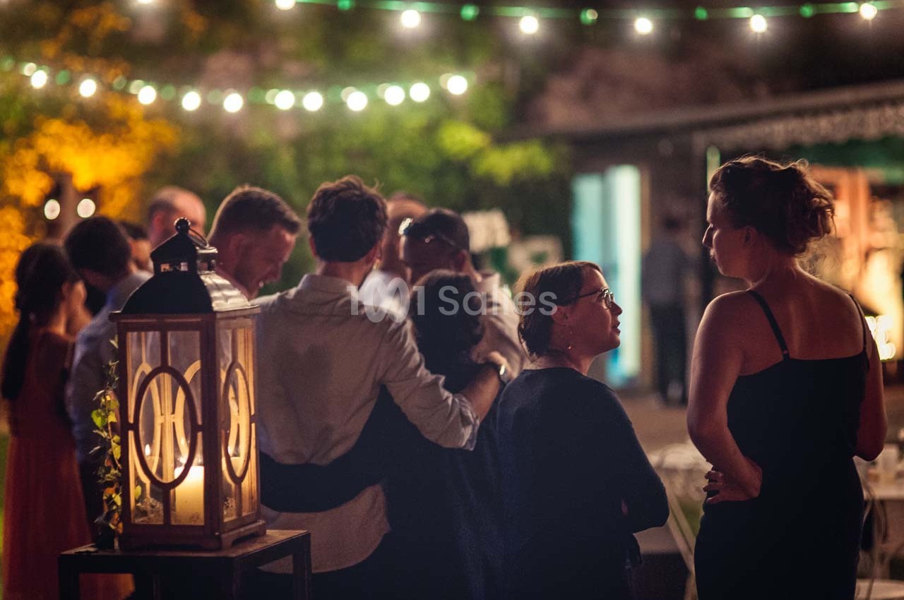 Groupe de personnes discutant dans un jardin éclairé par des guirlandes lumineuses, ambiance chaleureuse en soirée.