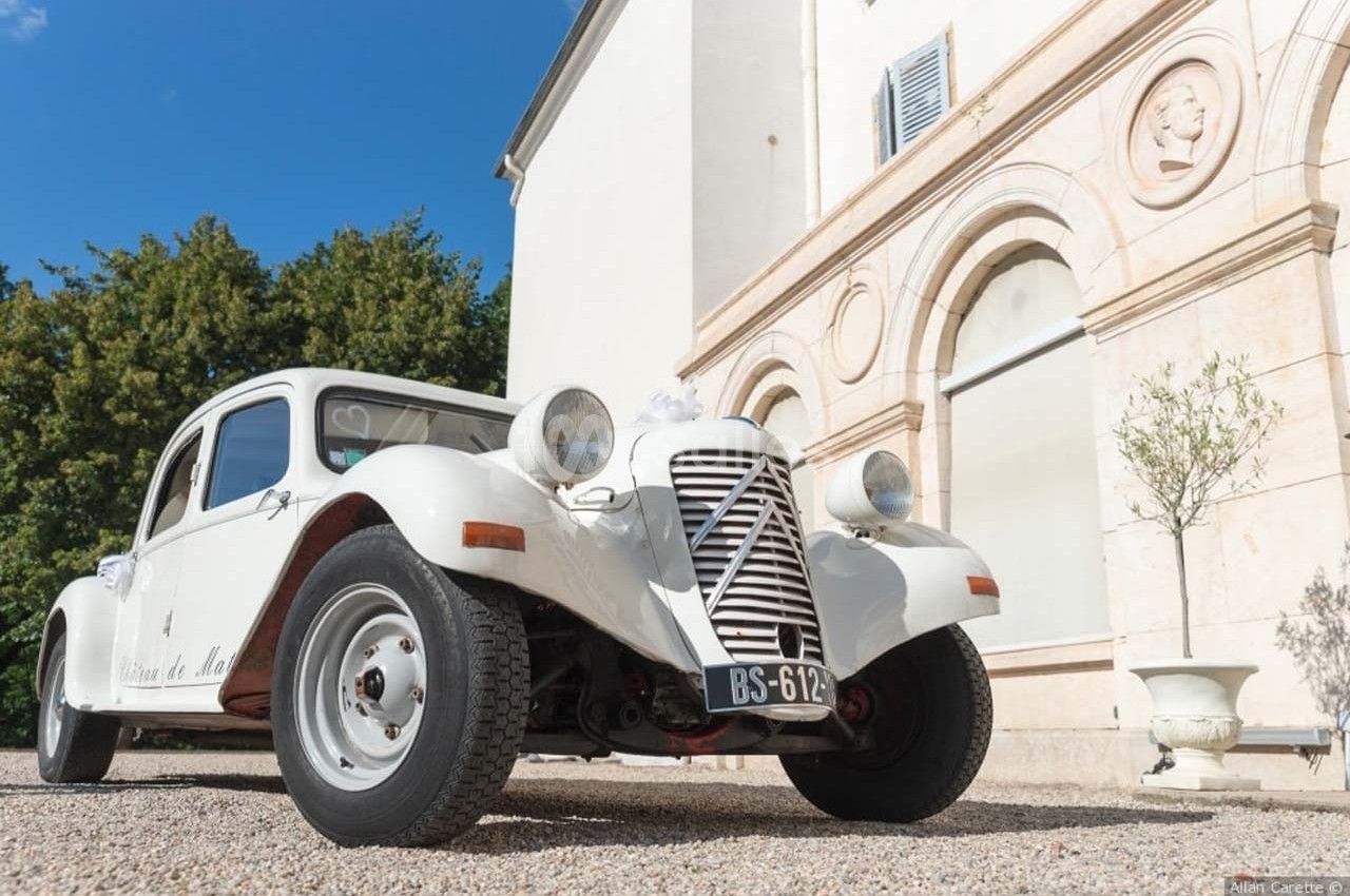 Voiture ancienne blanche stationnée devant un bâtiment clair avec des arches et des arbres en arrière-plan.