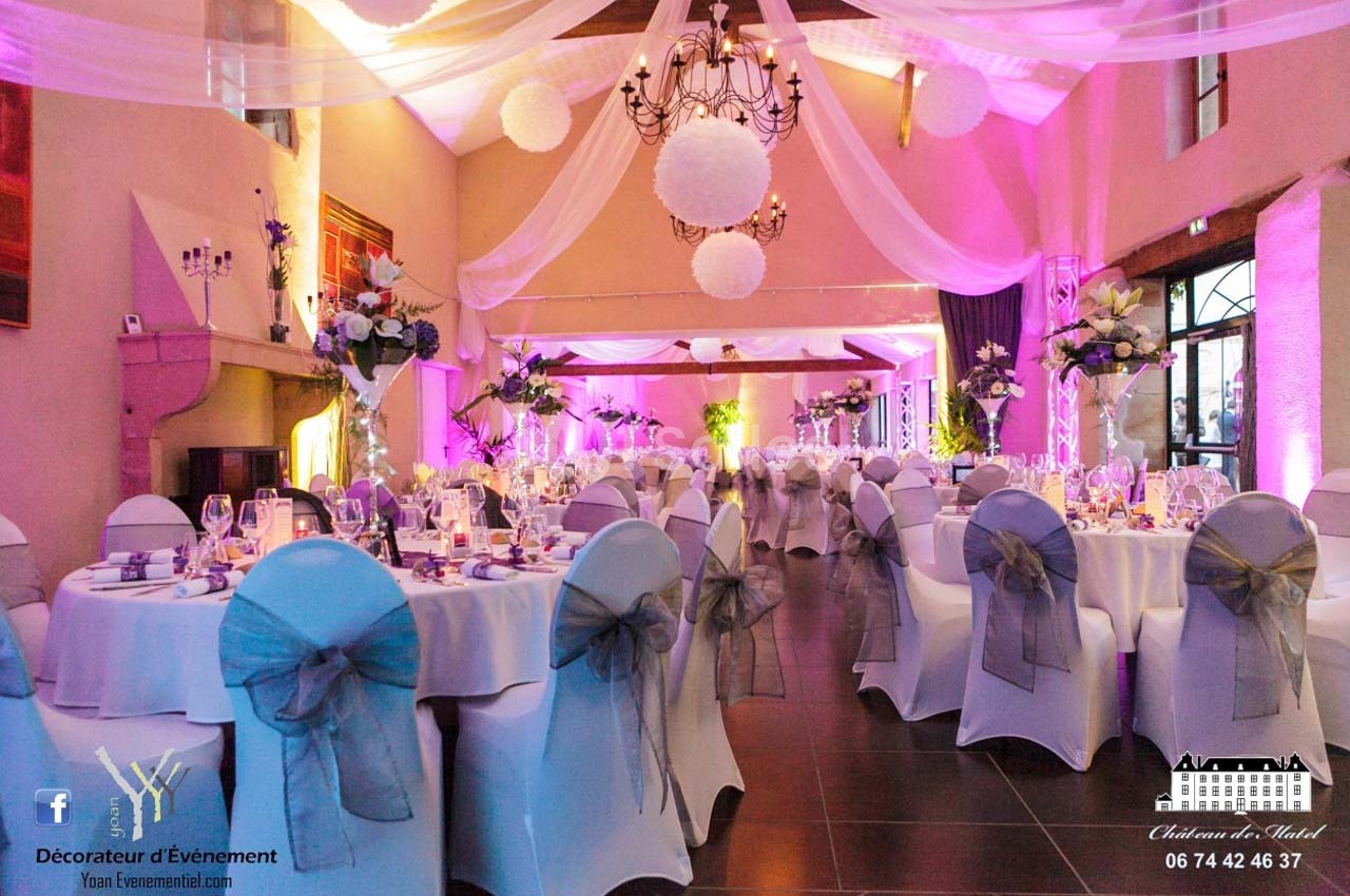 Salle de réception décorée avec des tables rondes, nappes blanches, chaises ornées de nœuds gris et éclairage rose.