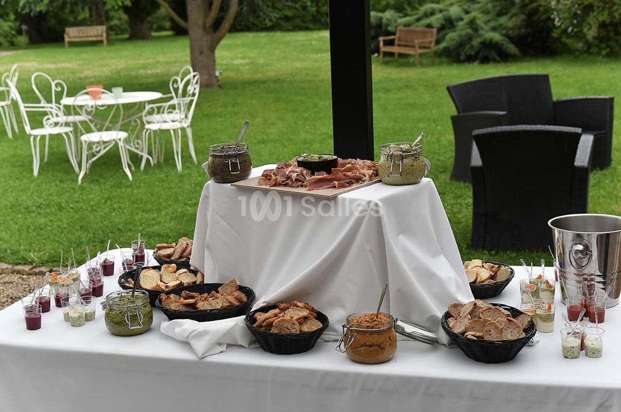 Table de buffet en extérieur avec pain, pâtés, condiments et verrines, dans un jardin avec mobilier de jardin en arrière…