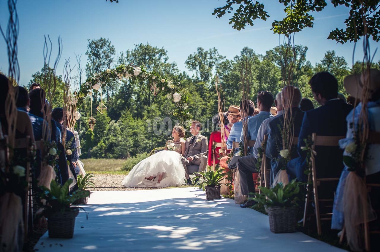 Un couple assis sous une arche fleurie lors d'une cérémonie de mariage en plein air, entouré d'invités.