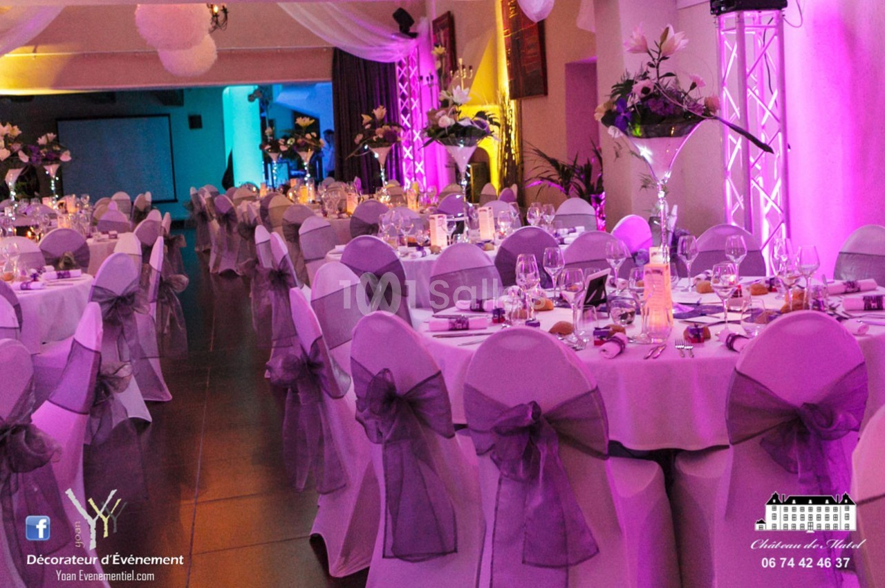 Salle de réception décorée avec des tables rondes, nappes blanches, chaises ornées de nœuds violets et éclairage coloré.