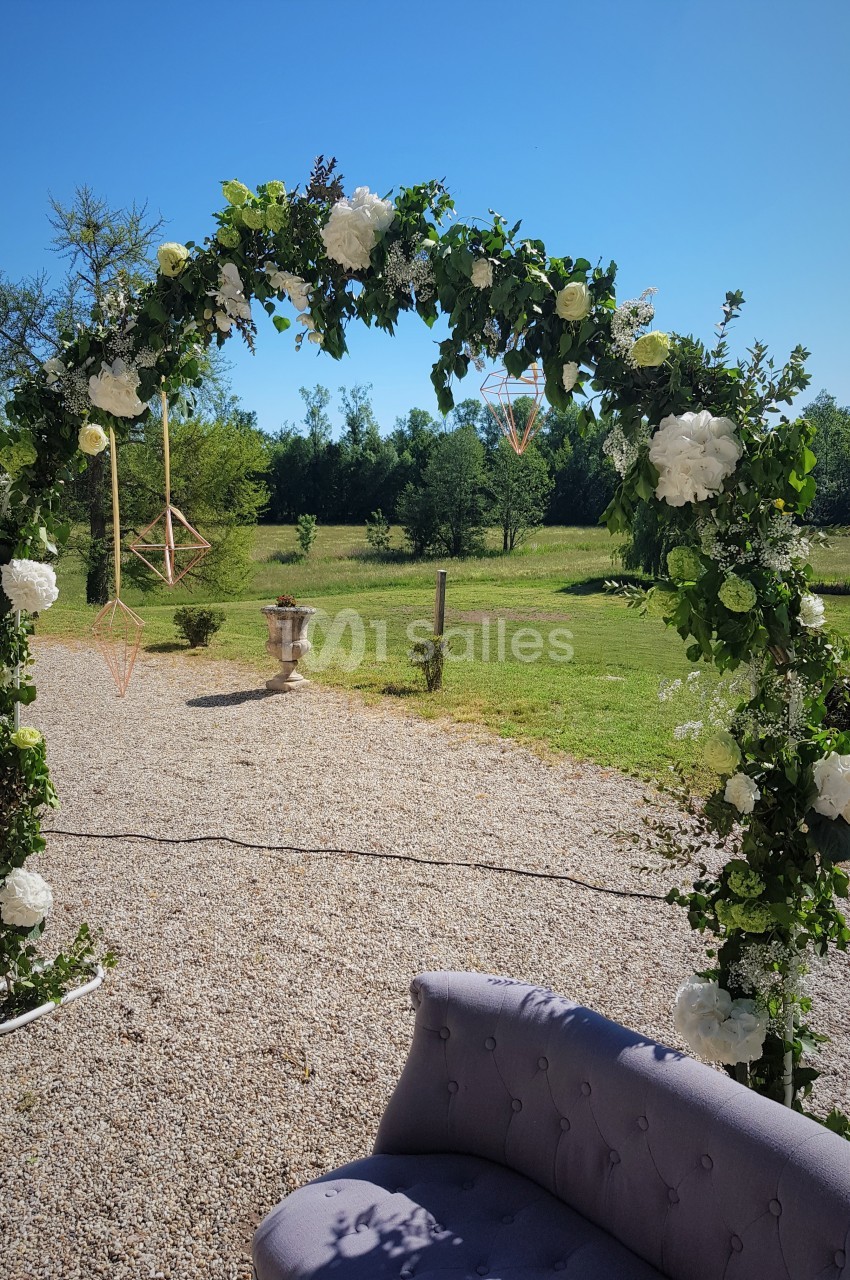 Arche de mariage décorée de fleurs blanches et feuillage, installée sur un chemin gravillonné avec vue sur un jardin.