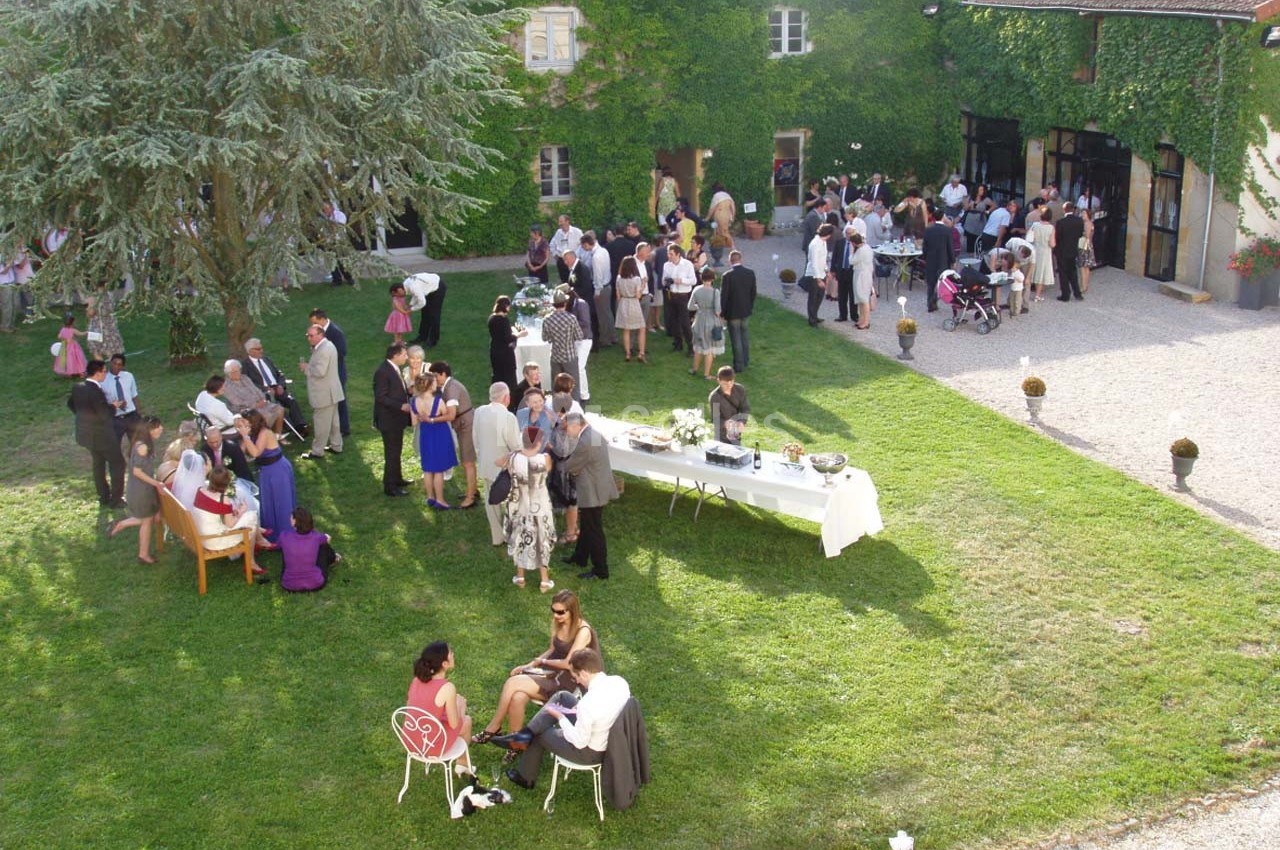 Groupe de personnes rassemblées dans un jardin pour une réception, avec tables et bâtiments en arrière-plan.