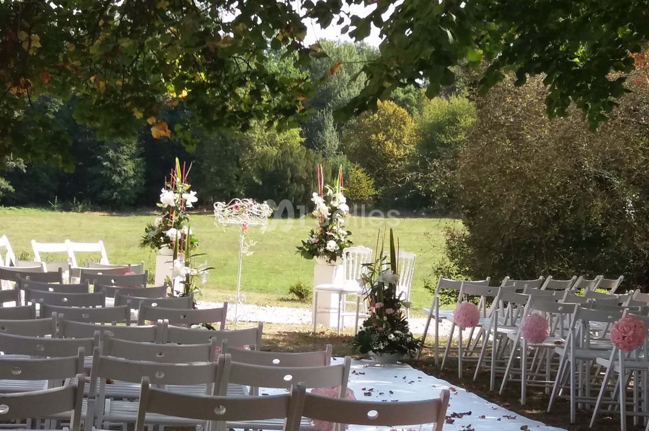 Chaises blanches alignées en extérieur, décorées de fleurs, face à une arche florale dans un cadre verdoyant.