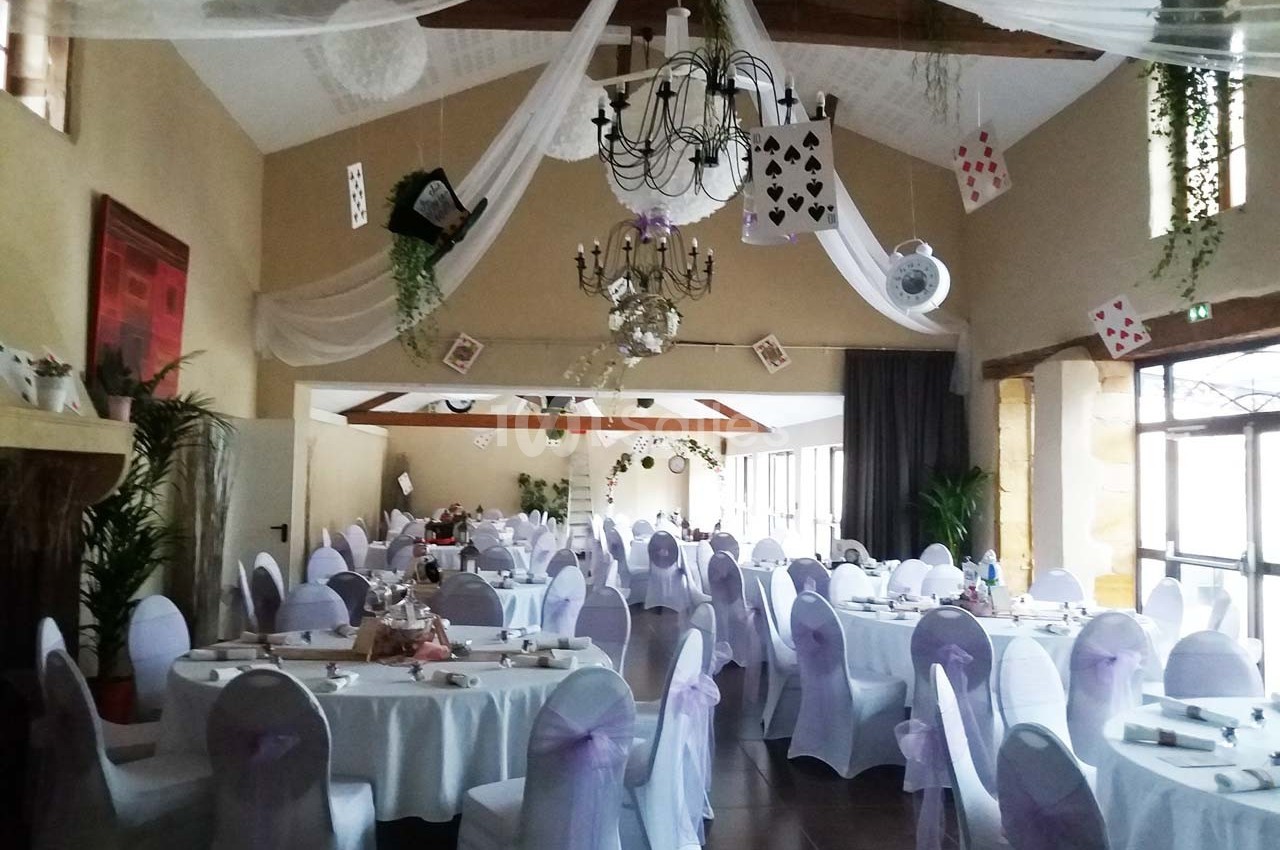 Salle de réception décorée avec des nappes blanches, des chaises ornées de rubans violets et des cartes suspendues.