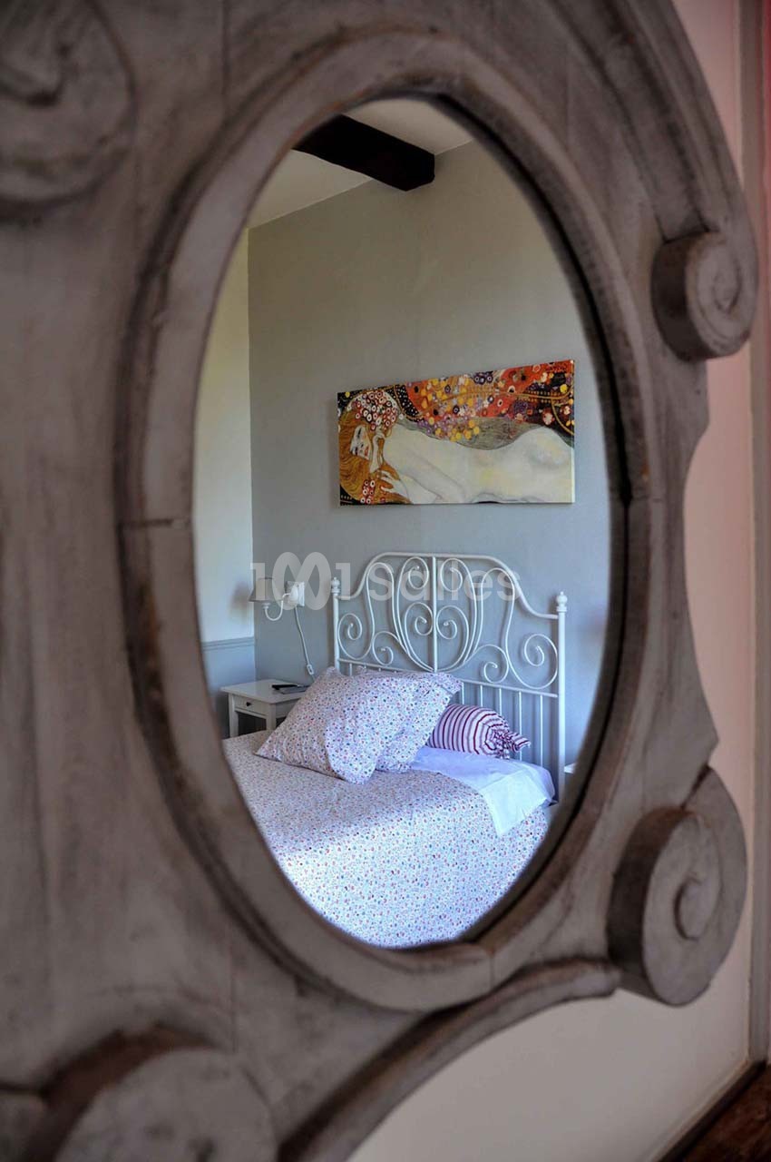 Reflet d'un lit avec une tête de lit blanche et une peinture colorée en arrière-plan, vu à travers un miroir ovale.